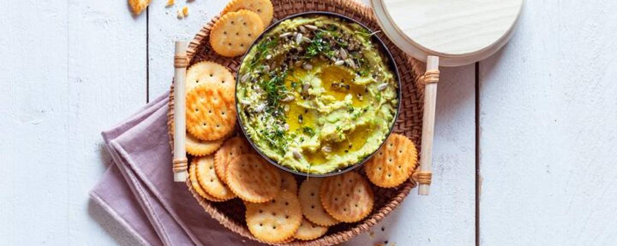 Avocado-Dip in Schüssel mit Cracker auf Teller mit rosanem Geschirrhandtuch