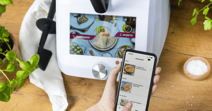 Monsieur Cuisine im Hintergrund, im Vordergrund eine Hand, die ein Handy hält. Auf dem Display ist die ZauberMix App mit dem Wochenplaner.