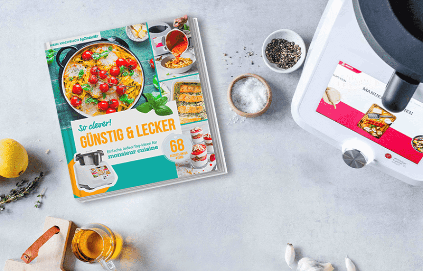 Neues ZauberMix-Kochbuch "Günstig & Lecker" liegen neben dem Monsieur Cuisine und umringt von Zutaten auf einer Arbeitsplatte